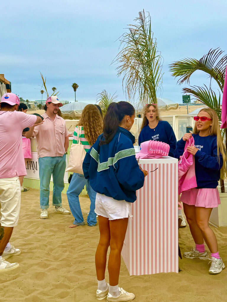 Three girls wearing Hampton Water sweats around a stack of pink baseball caps